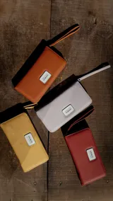 Billetera larga con cierre perimetral, confeccionada en material sintético texturizado color mostaza. Cuenta con correa de mano extraíble y placa metálica con logo de la marca en el frente.