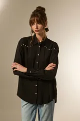 Camisa negra de viscosa con corte holgado, cuello clásico, cierre frontal de botones y detalle de tachas metálicas en la línea de los hombros.