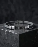Pulsera masculina con diseño de eslabón central de acero inoxidable con patrón geométrico griego, flanqueado por secciones de acero y correa de cuero gris.