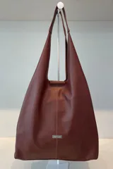 Bolso tipo tote de gran capacidad, confeccionado artesanalmente en cuero vacuno color marrón. Presenta un diseño minimalista con costura central vertical, fuelle lateral con cierre para ajustar su volumen y placa metálica con logo de la marca en el frente.