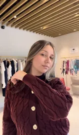 Cardigan de punto trenzado color beige, con cuello redondo, cierre frontal con botones dorados y mangas largas con puños acanalados.