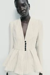 Blazer entallado de color crema con rayas verticales finas, cuello sin solapa y manga larga con hombreras. Presenta un diseño con bajo en evasé tipo peplum y cierre frontal con botones negros.