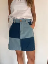 Minifalda de denim con diseño patchwork en tonos azul oscuro y celeste, tiro alto, bolsillos delanteros y cierre frontal con botón y cremallera.