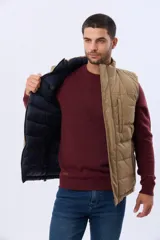 Chaleco reversible acolchado de corte recto, con cuello alto y cierre frontal de cremallera. Un lado es de color tostado con pespuntes horizontales y un bolsillo vertical con cierre en el pecho, mientras que el reverso es de color azul marino con diseño capitonado.