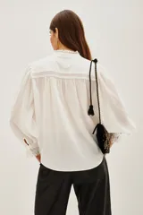 Camisa blanca de cuello alto con volados y encaje, mangas largas abullonadas con puños de encaje. Presenta detalles de encaje y pliegues verticales en el frente.