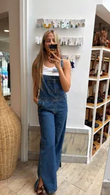 Enterito largo de denim negro con diseño de pierna ancha, bolsillo frontal en el peto y aberturas laterales en el bajo.