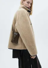 Chaqueta de efecto pelo en color beige, con diseño oversize y corte recto. Presenta cuello alto, cierre frontal con botones y detalles de ribetes en cuero sintético negro.