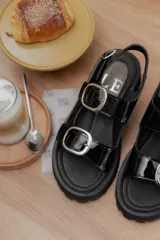 Sandalias de plataforma estilo chunky en color negro, confeccionadas en material sintético efecto charol. Presentan dos tiras ajustables con hebillas metálicas plateadas y una tira trasera con hebilla para mayor sujeción. Suela dentada de goma.