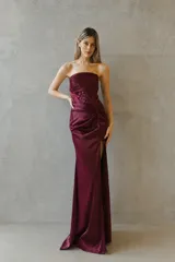 Vestido largo strapless de satén color bordó, con cuerpo estilo corsé estructurado y detalle de drapeado lateral que desemboca en una abertura frontal en la falda.