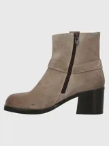 Bota corta de descarne color beige, con taco cuadrado y punta cuadrada. Presenta una correa con argolla metálica decorativa alrededor del tobillo y cierre lateral.