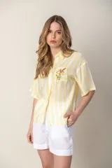 Camisa a rayas verticales amarillas y blancas, con cuello camisero, mangas cortas y bolsillo en el pecho con bordado de un pájaro.