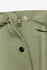 Chubasquero de acabado engomado en color verde militar, con capucha ajustable mediante cordones, cierre frontal con cremallera y botones a presión ocultos, y bolsillos laterales con solapa.
