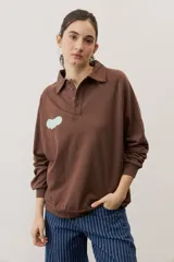 Buzo celeste de manga larga con cuello polo y cierre frontal de botones. Presenta un parche marrón bordado en el pecho.