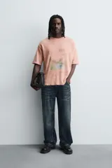 Camiseta de punto de algodón con corte boxy fit, cuello redondo y manga corta. Presenta un estampado frontal con diseño de paisaje y texto en tonos deslavados. La prenda tiene un efecto lavado que le da un aspecto envejecido e irregular.