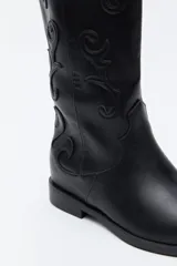 Bota de caña alta estilo western confeccionada en cuero sintético, con bordados decorativos en relieve a lo largo de la caña y puntera afinada.