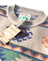 Buzo tejido de punto con diseño geométrico inspirado en motivos andinos, en tonos beige, verde, azul y naranja. Presenta cuello redondo, puños y ruedo acanalados, y etiqueta de marca en el frente.