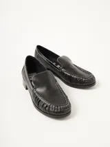 Mocasines de cuero negro con costuras pespunteadas en hilo blanco a contraste, diseño fruncido en la puntera, talón plano con pequeño tacón y apliques sutiles en charol.