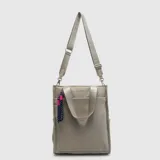 Bolso tipo tote Bubba modelo Ontario Regular, color gris claro, confeccionado en cuero sintético. Cuenta con compartimento principal con cierre, asas de mano y una correa larga ajustable con logo impreso. Incluye un llavero decorativo de cordón trenzado en colores fucsia y azul.
