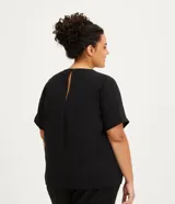 Blusa de manga corta color beige, confeccionada en tela texturizada. Presenta cuello redondo con detalle de pespuntes y cierre trasero con botón en forma de lágrima.