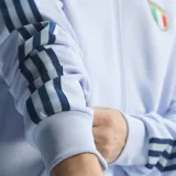 Campera deportiva Adidas de la Selección de Italia, color celeste, con cierre frontal y las clásicas tres tiras en azul marino en las mangas. Cuenta con bolsillos laterales, cuello alto y los escudos de Adidas y de la federación italiana bordados en el pecho.