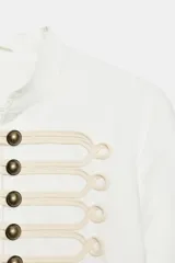 Chaqueta entallada de color blanco con cuello redondo y manga larga. Presenta un diseño de estilo militar con cierre frontal mediante alamares decorativos y botones metálicos.