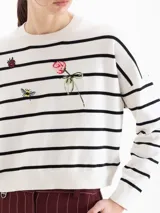 Sweater de tejido liviano color blanco con rayas horizontales negras, escote redondo, mangas largas y hombros caídos. Cuenta con pequeños bordados decorativos en el frente que representan una abeja, una flor y una frutilla.