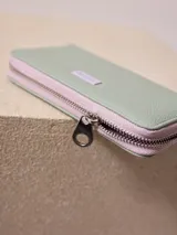 Billetera rectangular color verde agua, con cierre perimetral de cremallera rosa y aplique metálico con el logo de la marca. Interior con dos compartimentos.