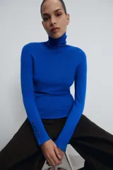 Jersey de punto liso en color beige, con cuello alto y manga larga con detalle de botones metálicos en los puños.