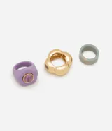 Set de tres anillos con diseños variados: uno de resina color lila con detalle central circular, uno de metal dorado con forma de flor y uno de resina con efecto marmolado en tonos azulados.