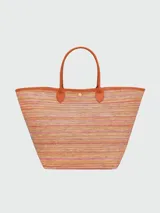 Bolso tipo cesta de hombro Longchamp modelo Le Pliage, confeccionado en tejido de paja con rayas multicolor. Presenta detalles en cuero color naranja, incluyendo el bolsillo frontal con el logo grabado y las asas. Incluye un colgante decorativo con cuentas de madera.