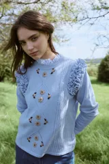 Sweater de punto color celeste, con diseño de rombos calados y bordados florales artesanales en el frente. Presenta volados decorativos en los hombros y cuello redondo.