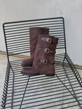 Botas de caña alta de gamuza marrón con puntera cuadrada, suela plana y tres tiras laterales con hebillas metálicas.