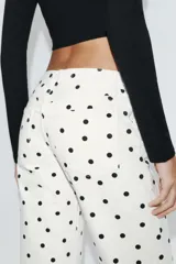 Jeans de tiro bajo con diseño de pierna ancha y corte largo. Presenta un estampado de lunares negros sobre fondo blanco, cierre frontal con cremallera y botón metálico, y cinco bolsillos.