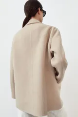Abrigo corto de Zara confeccionado en hilatura con lana, de color beige. Presenta cuello solapa, manga larga, cierre frontal con botones negros y bolsillos de plastrón en el delantero.