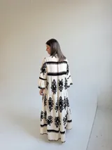 Vestido largo color marfil con estampado ornamental en color negro, escote en V con botones frontales, mangas largas amplias y diseño escalonado en la falda.