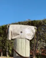 Bandolera de cuero color plata macchiato, con acabado brillante y correa ajustable marrón. Presenta un bolsillo frontal con solapa y cierre de broche, y un detalle de flecos en el lateral.