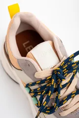 Zapatilla urbana de diseño chunky, con paneles combinados en tonos beige, arena y gris. Presenta cordones jaspeados en azul y amarillo, tirador trasero amarillo y suela de plataforma dentada en color crema.
