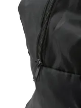 Bolso de viaje tipo duffle color negro, confeccionado en material sintético ligero. Presenta correas de hombro en color gris claro que recorren el cuerpo del bolso, un bolsillo frontal con cierre y una etiqueta decorativa circular en la parte inferior.