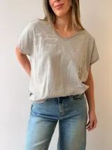 Remera gris claro de corte relajado, con escote en V, mangas cortas y estampa con texto manuscrito. Cuenta con un bolsillo frontal decorado con apliques de strass y tachas metálicas, además de detalles desflecados.