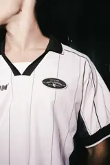 Camiseta de estilo deportivo con cuello polo, confeccionada en tela técnica de secado rápido. Presenta un diseño a rayas verticales en color rosa con paneles laterales en malla negra, mangas cortas con bordes negros y detalles gráficos estampados en el frente.