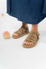Sandalia estilo gladiadora de color beige, confeccionada en material gamuzado sintético. Presenta múltiples tiras anchas que sujetan el pie, ajuste lateral con hebilla metálica y suela plana antideslizante de color marrón.