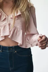 Blusa corta color rosa pálido con tejido suave y leve brillo. Presenta un escote en V pronunciado adornado con un ribete ancho de volados que continúa en el bajo de la prenda. Tiene mangas largas con volumen y puños con volado.