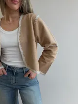 Chaqueta de efecto gamuza color tostado, con diseño abierto sin cierres y detalles de costuras artesanales tipo crochet en color blanco en los bordes, el frente, el cuello y las mangas.