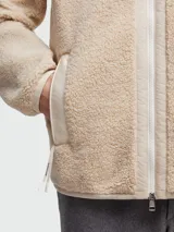 Buzo de polar tipo teddy en color beige, con cierre frontal completo, cuello alto y bolsillo en el pecho con solapa y logo de la marca. Presenta detalles en tela técnica al tono en los bordes de los bolsillos, cierre y cuello.