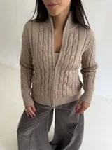 Sweater tipo campera de punto color beige, con cierre frontal completo y diseño de trenzas verticales. Presenta cuello alto y puños acanalados.