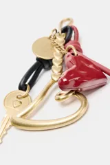 Llavero accesorio con clip metálico, que incluye una llave dorada, un dije de corazón grande de metal dorado, un dije de corazón pequeño esmaltado rojo, un dije redondo dorado, una cadena con dijes de corazón pequeños dorados, y tiras de cuero anudadas en rojo y negro.