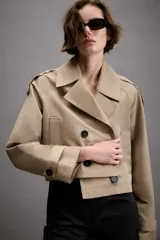 Trench corto de color beige con cuello de solapa, cierre frontal cruzado con botones y mangas largas con trabillas ajustables en los puños. Presenta un diseño estructurado con hombreras y bolsillos delanteros de vivo.