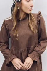 Vestido corto de gamuza color marrón, con diseño de corte evasé, volados en los hombros y mangas largas con puños elásticos fruncidos. Presenta un cuello alto y una falda con un volado en el ruedo.