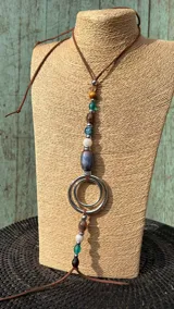 Collar tipo colgante con tira de cuero marrón, cuentas multicolores de vidrio y piedra, separadores metálicos y aros concéntricos de metal plateado.