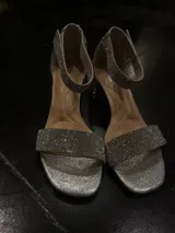 Sandalia de fiesta con taco cuadrado bajo, confeccionada en material plateado con acabado brillante. Presenta una tira frontal ancha y una pulsera al tobillo, ambas completamente cubiertas de strass.